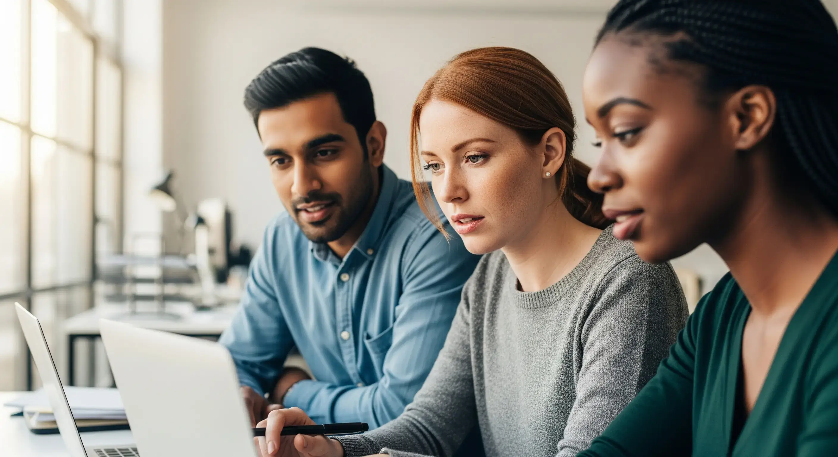 Eine diverse Gruppe von drei Fachleuten arbeitet in einem hellen, modernen Büro konzentriert an einem Laptop zusammen.