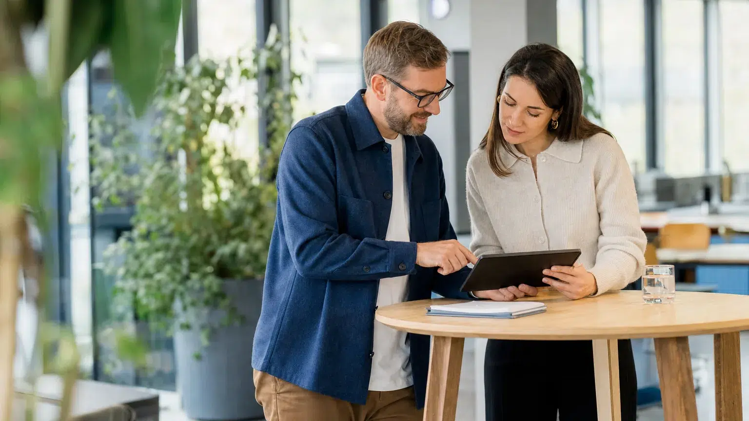 Ein Mann und eine Frau besprechen gemeinsam Unterlagen auf einem Tablet an einem Stehtisch im Büro